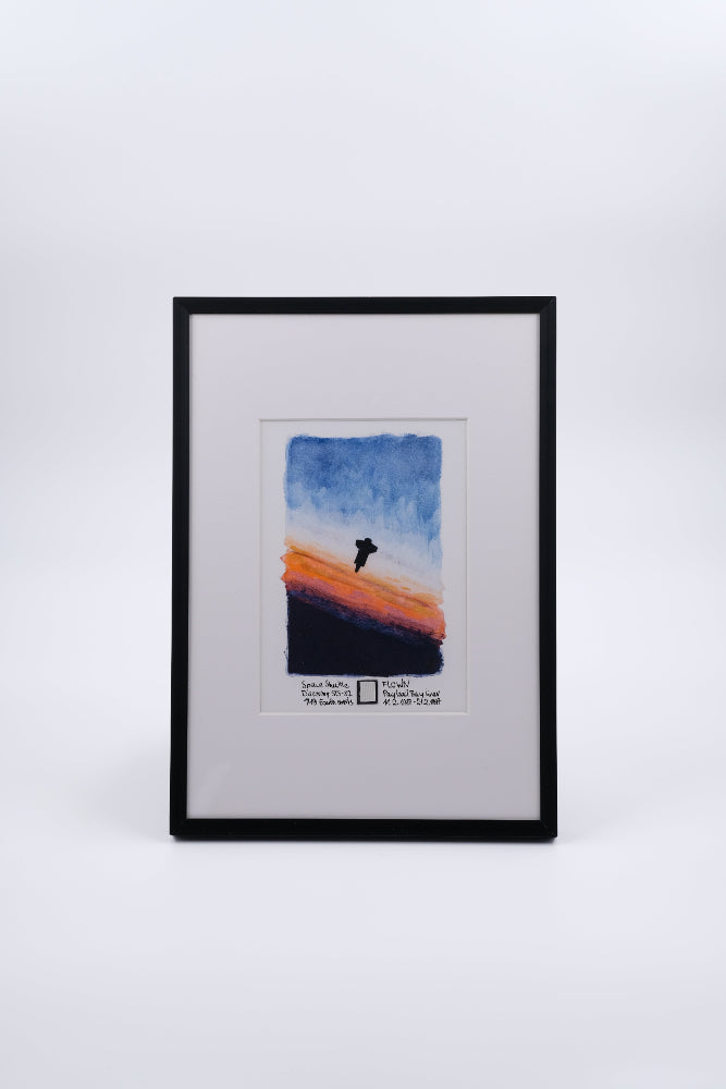 Framed watercolor artwork showing a space shuttle silhouette against an orbital sunrise gradient, with a small embedded flown payload bay liner fragment in the mat.