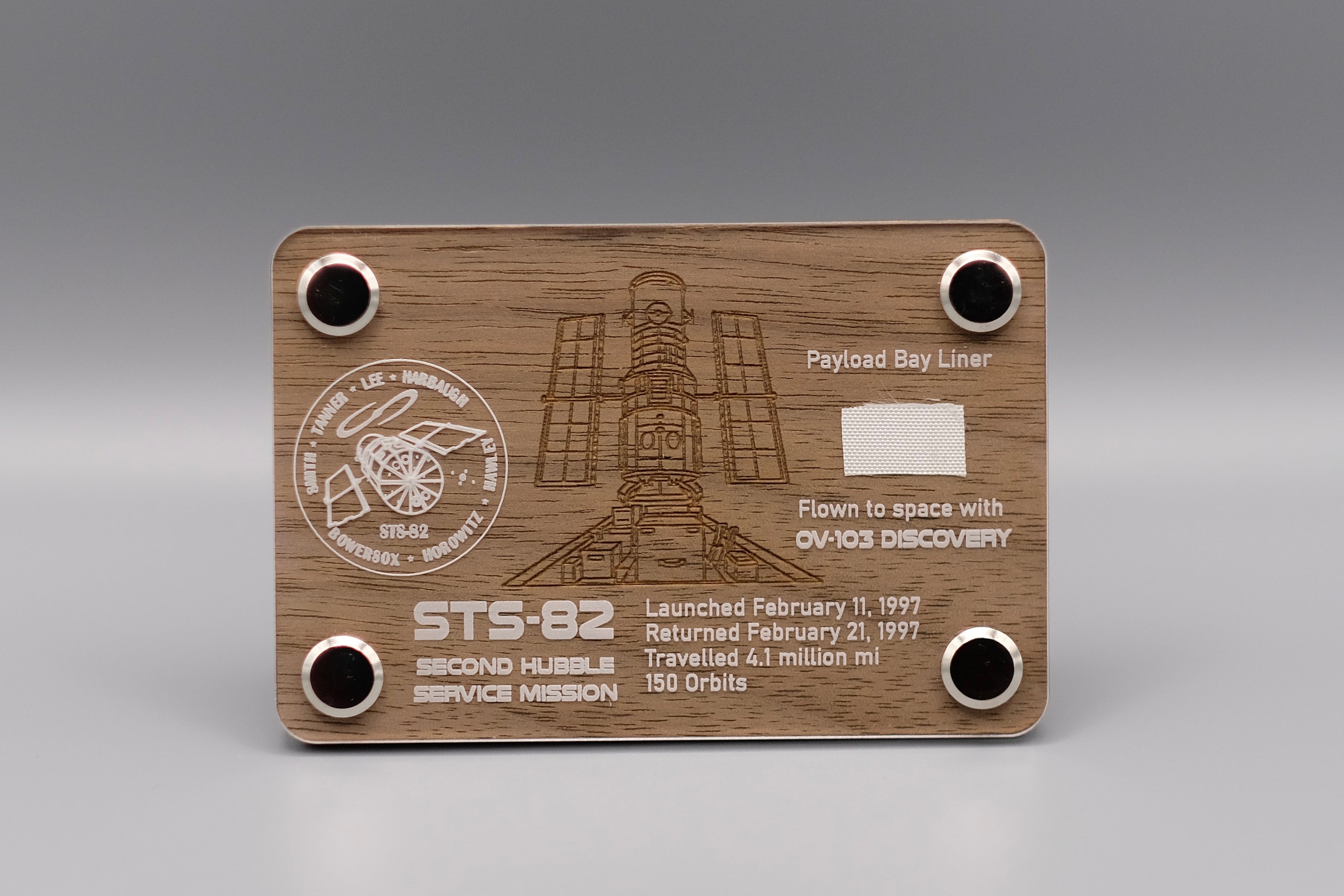 Wooden display with authentic flown Payload Bay Liner fragment from Space Shuttle Discovery STS-82 mission, engraved with Hubble Space Telescope servicing details.