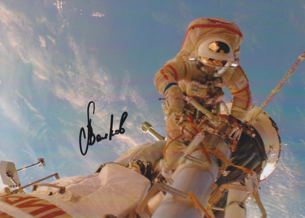 Color photograph of a cosmonaut (Volkov)conducting an EVA above Earth with a visible handwritten signature on the image.
