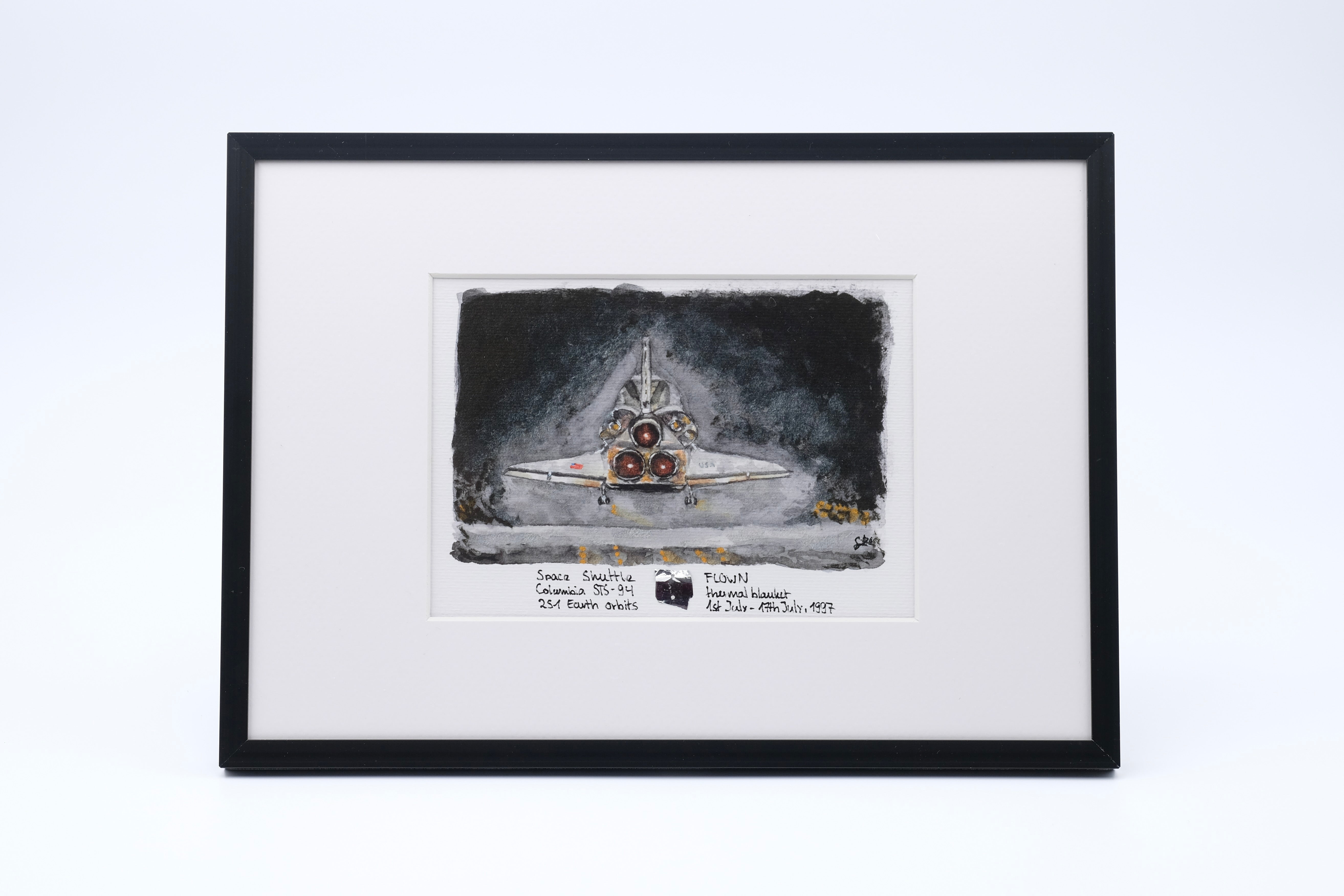 Framed watercolor artwork showing the rear view of Space Shuttle Atlantis with its three main engines against a dark background, featuring a small embedded flown solar blanket fragment in the mat.