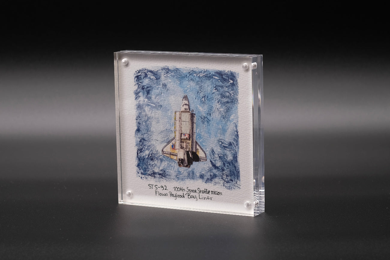 Acrylic display with watercolor artwork of Space Shuttle Discovery and a flown STS-82 payload bay liner fragment inside.