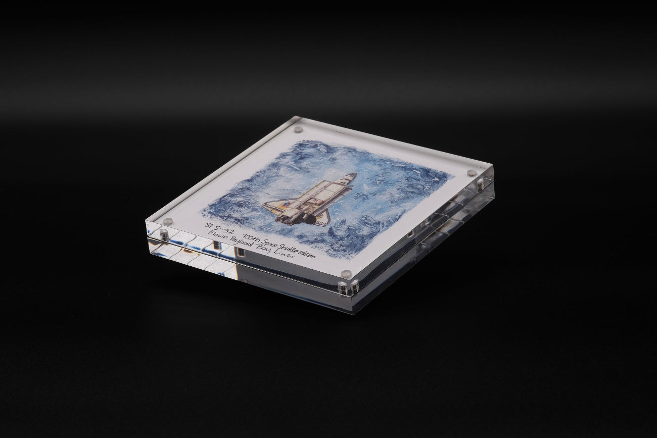 Acrylic display with watercolor artwork of Space Shuttle Discovery and a flown STS-82 payload bay liner fragment inside.